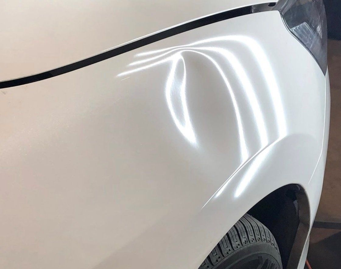 White car fender with a prominent dent, reflecting overhead lights.