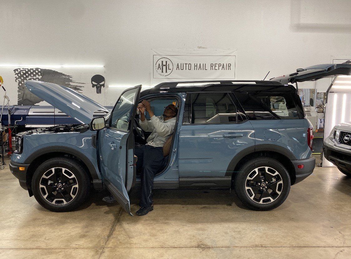 Man inside a blue Ford Bronco with open doors and hood, in a repair shop.