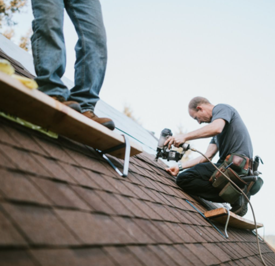 Two roofers installing asphalt shingles on a sloped roof using a pneumatic nail gun.