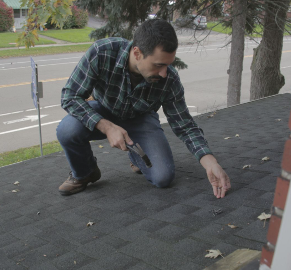 A person in a plaid shirt kneels on a residential roof, using a hammer to repair asphalt shingles.