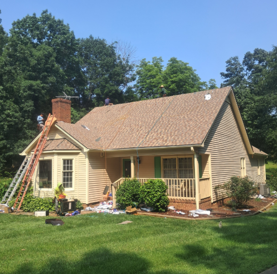 Workers replacing the shingles on the brown roof of a single-story beige house with a chimney and front porch.