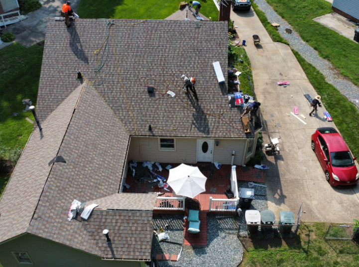 A high-angle view shows roofers replacing shingles on a house, with debris on the deck and a red car parked in the driveway.