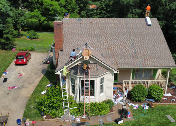 Roofers work on the shingled roof of a suburban house, using ladders and tools, with materials scattered on the lawn below.