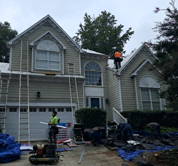 Roofers in high-visibility vests work on a two-story beige house, replacing shingles with tools and ladders present.