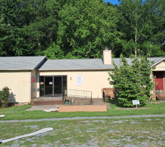 Single-story tan building with a grey roof, a ramp to the entrance, and a red door, set against a wooded backdrop.