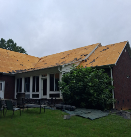 A red brick house with a partially removed shingle roof revealing the wooden sheathing, set against a cloudy sky.