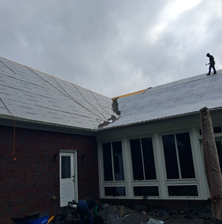 Workers repair a residential roof covered in white underlayment above a brick house with large windows.