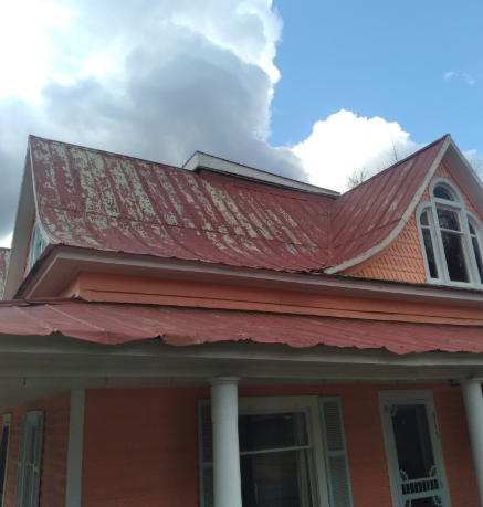 The corner of a peach-colored house with a weathered red metal roof against a bright blue, cloudy sky.