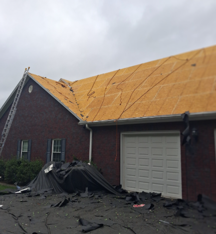 A residential brick home with its roof shingles stripped, exposing the plywood sheathing, with debris on the driveway.
