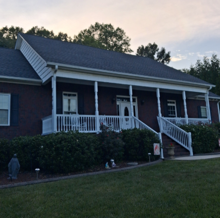 A dark brick ranch-style house with a white wraparound porch, stairs leading to the yard, and surrounding landscaping.