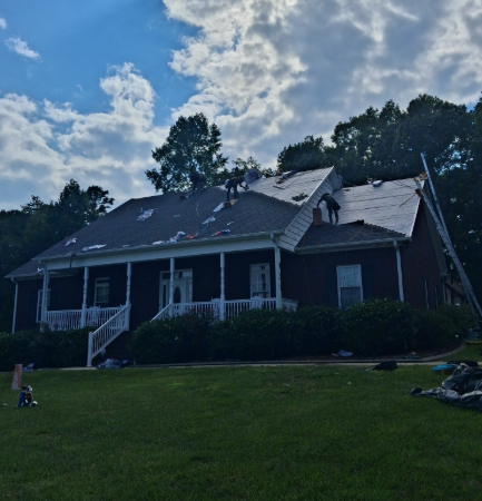 Workers perform roof repairs on a dark-sided house with a front porch under a partly cloudy sky.
