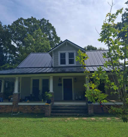 A single-story gray house with a dark metal roof, a front porch, and a dormer window, surrounded by trees and a lawn.
