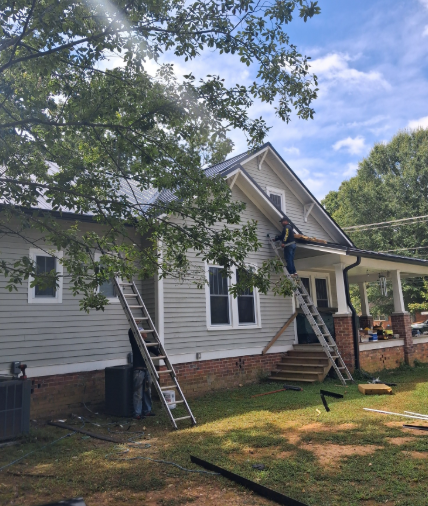 Two workers on extension ladders install a metal roof on a light gray house with a brick foundation and front porch.