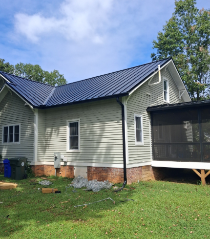 A light-colored house with a dark metal roof and an attached screened-in porch, situated on a lawn under a blue sky.