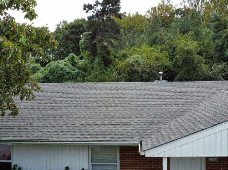 The gray shingled roof of a brick house is set against a dense background of green trees.