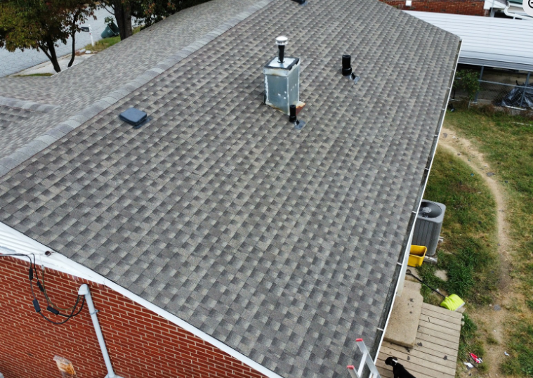 A high-angle view of a gray shingled roof on a brick house, featuring a chimney, vent pipes, and a suburban backyard.