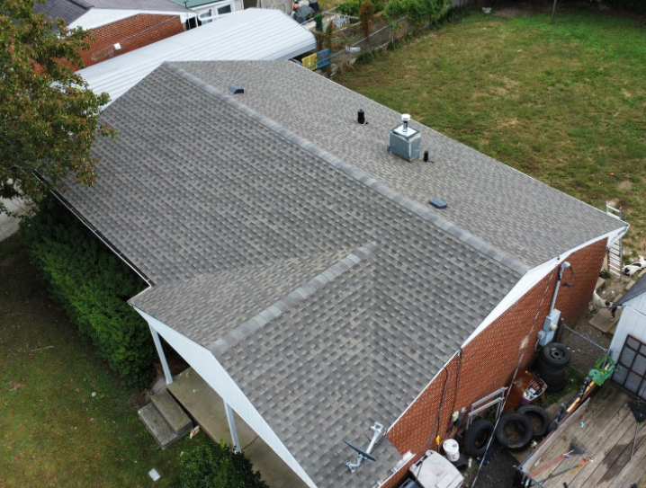 An aerial view of a gray-shingled roof on a brick house, featuring a covered porch and several roof vents.