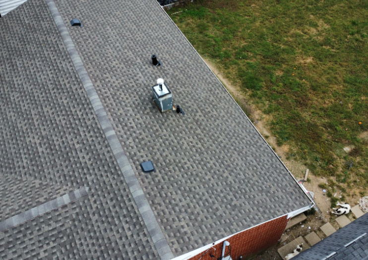 An aerial view of a gray shingled roof with several vents and a chimney, bordering a patch of grass.