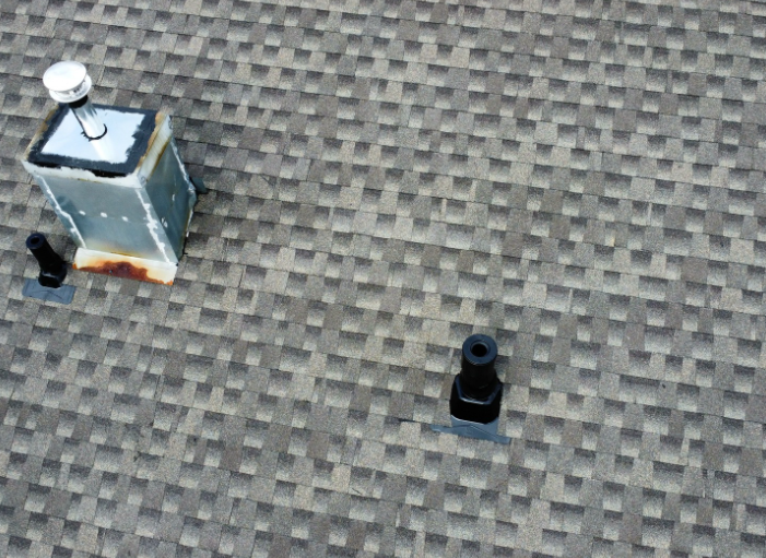 A metal chimney vent and two black plastic conduits protruding from a textured gray shingle roof.