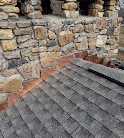 A stone chimney with copper roof flashing overlapping the shingles on an angled roof.