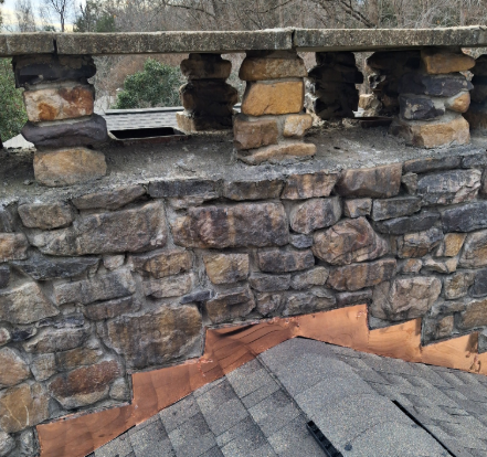 Stone chimney with an open-air top and copper flashing installed at the base where it meets a shingled roof.
