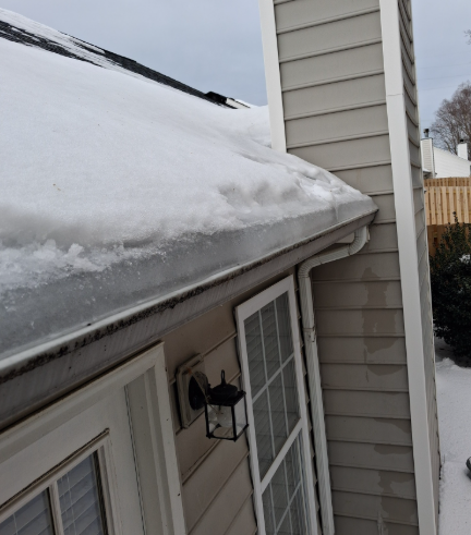 A thick layer of snow sits on a roof gutter next to a beige-sided chimney and house wall on an overcast day.