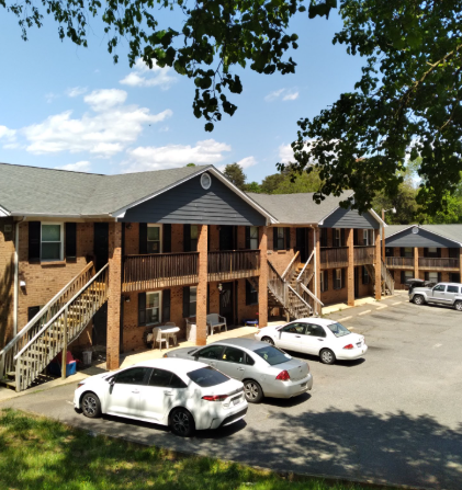 A multi-story brick apartment building with wooden stairs, dark blue accents, and cars parked in a sunny lot.
