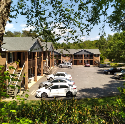 A two-story apartment complex with brick siding and a parking lot containing several cars on a sunny day.