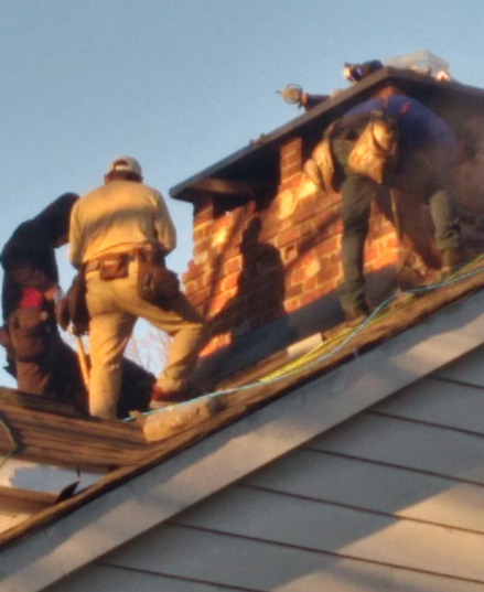 Three workers repair a brick chimney on a sloped shingled roof during sunset.