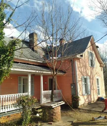 A light pink, two-story house with a porch and brick well in the front yard, where workers are performing roof repairs.