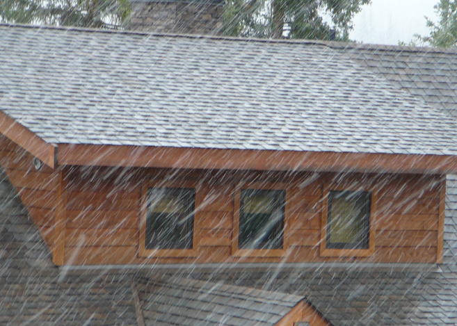 Heavy rain falls on a grey shingled roof and the wood-paneled exterior of a building with three windows.