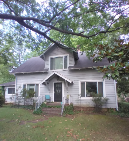 A light gray, two-story house with a prominent center dormer and a covered entryway sits on a grassy lot under large trees.