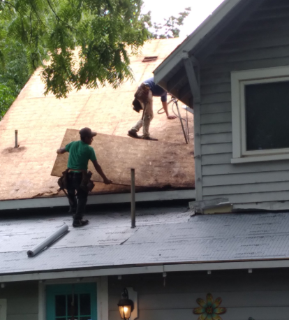 Two workers on a residential roof installing plywood sheathing over a partially shingled surface.