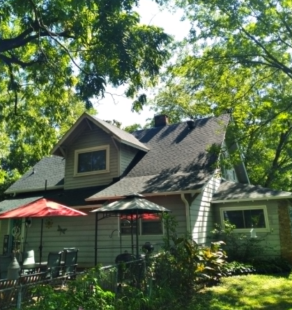 A two-story light-gray house with a dark roof nestled among large, leafy trees, with a red patio umbrella in the yard.