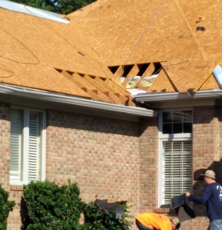 Two workers install plywood sheathing on a roof corner of a brick house with visible rafters.