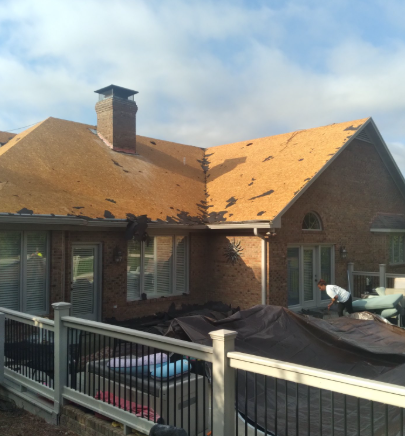 A worker on a residential deck prepares to repair a brown shingled roof under a partially cloudy sky.
