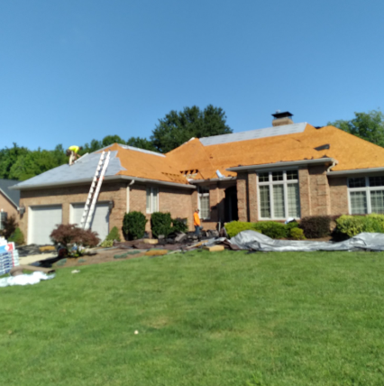 A brick house under construction with a stripped roof, a ladder, and workers outside on a sunny day.