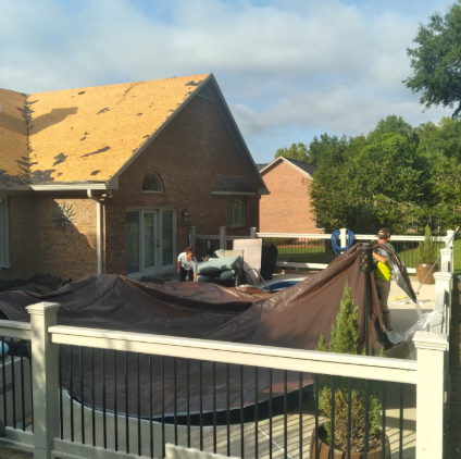 Two people unfold a large brown pool cover over an outdoor pool deck next to a house undergoing roof repairs.