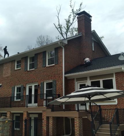 Two workers perform repairs on the roof of a two-story brick house with a deck and umbrella in the backyard.