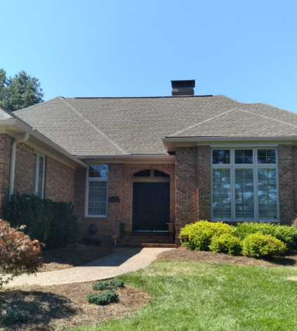 A single-story brick house with a dark front door, a light gray roof, and a green lawn under a clear blue sky.