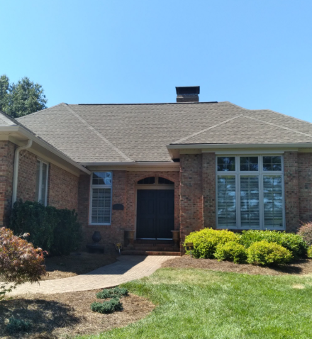 A brick house with a dark double-door entrance, grey-shingled roof, a chimney, and green shrubs under a clear blue sky.