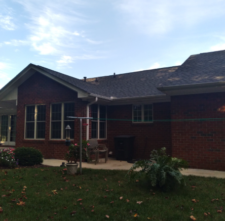 A single-story red brick house with a dark shingled roof, windows, and a paved patio area in a grassy yard.