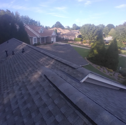 A high-angle view of a grey shingled residential roof, with nearby suburban homes and green trees in the background.