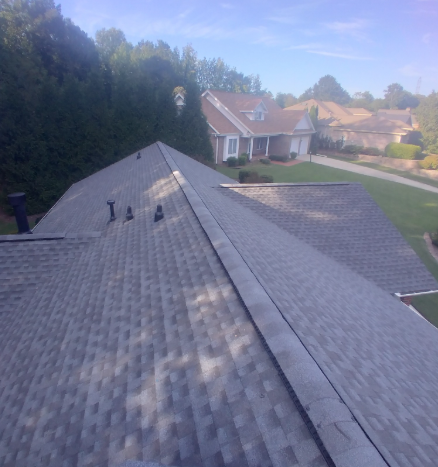 A high-angle view of a gray asphalt shingle roof on a sunny day, with a residential street and houses in the distance.