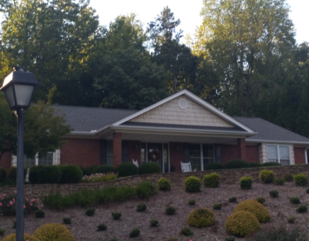 A brick house with a gabled roof and landscaped yard sits on a hillside beneath trees, with a street lamp in the foreground.