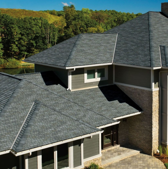 An exterior view of a multistory home with gray shingled roofs, dark siding, stone accents, and nearby trees.
