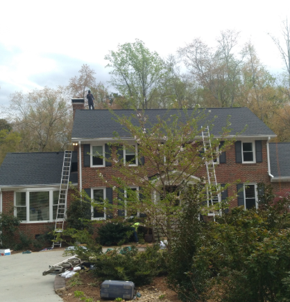 A person stands on the roof of a two-story brick house while workers install a new dark shingle roof using two ladders.