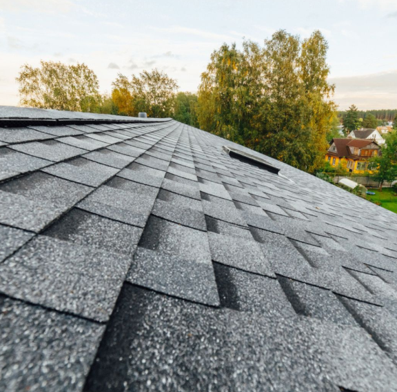 A close-up, low-angle view of a gray asphalt shingle roof, with blurred trees and houses in the background.