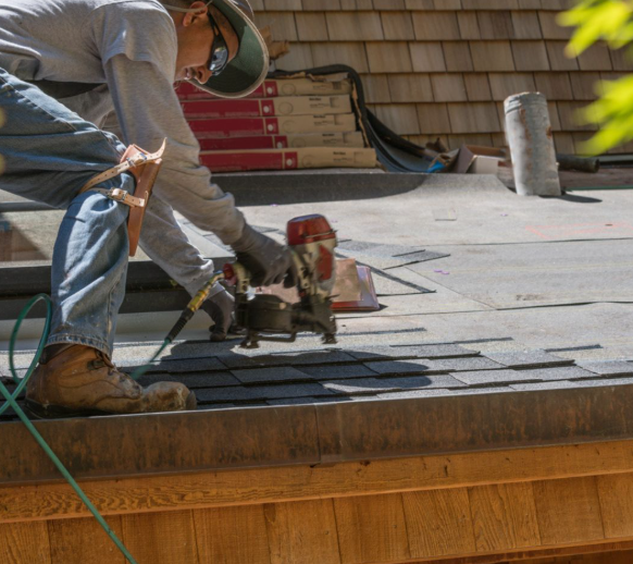 A worker in a hat, long sleeves, and jeans uses a pneumatic nail gun to install dark shingles on a residential roof.