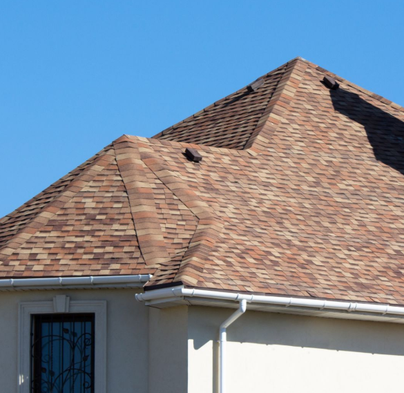 A close-up of a house roof with brown shingles, white gutters, and a blue sky background.